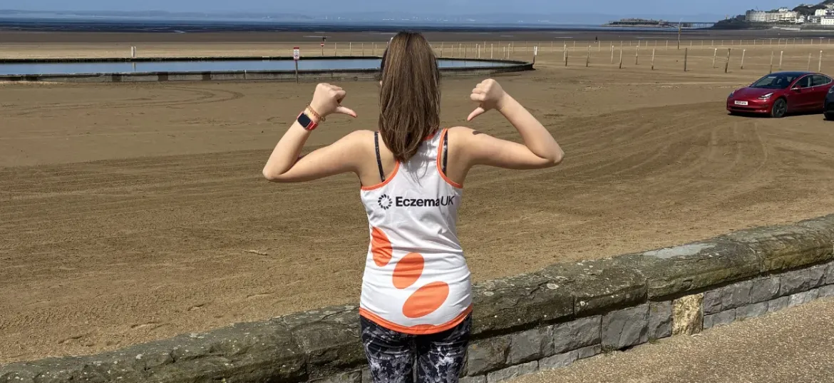 Hannah posing, in her Eczema UK running vest in front of a beach