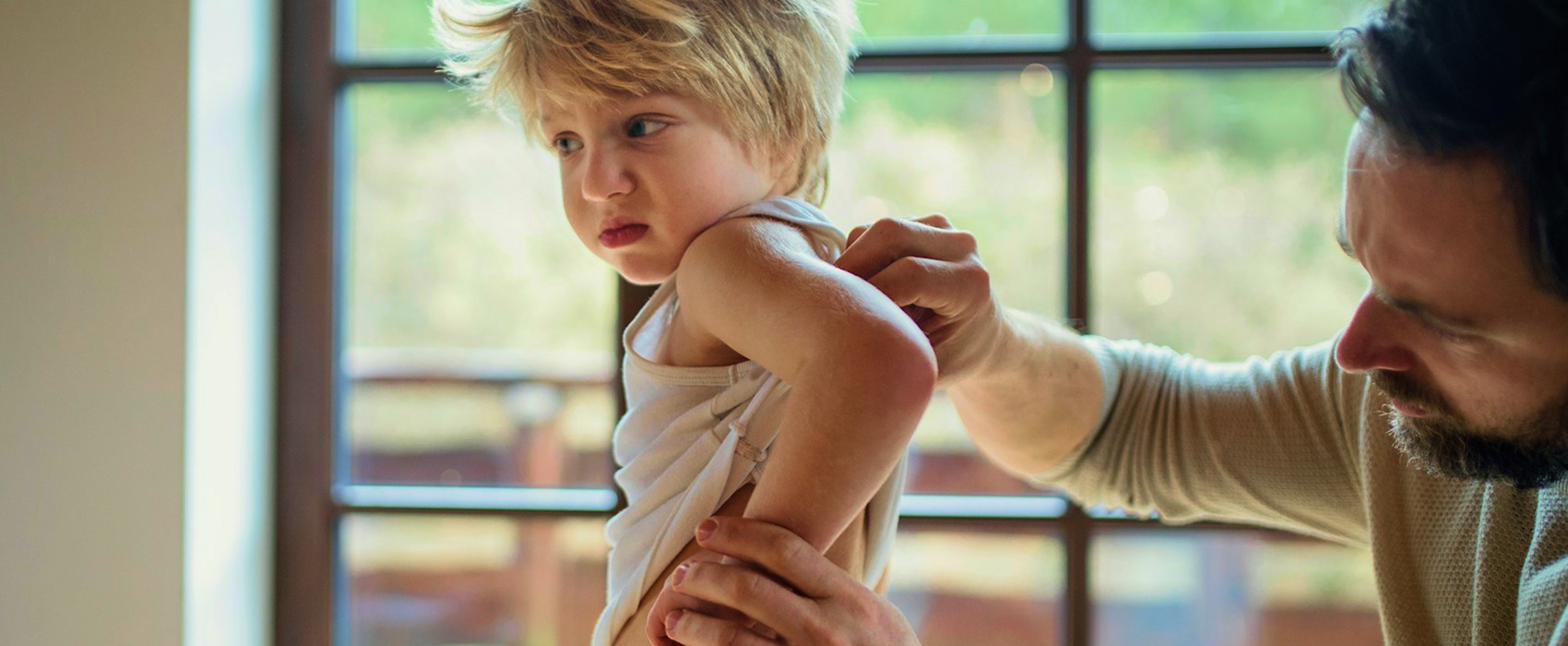A child is facing away from his father with a distressed look on his face as the father closely checks his sons back for eczema